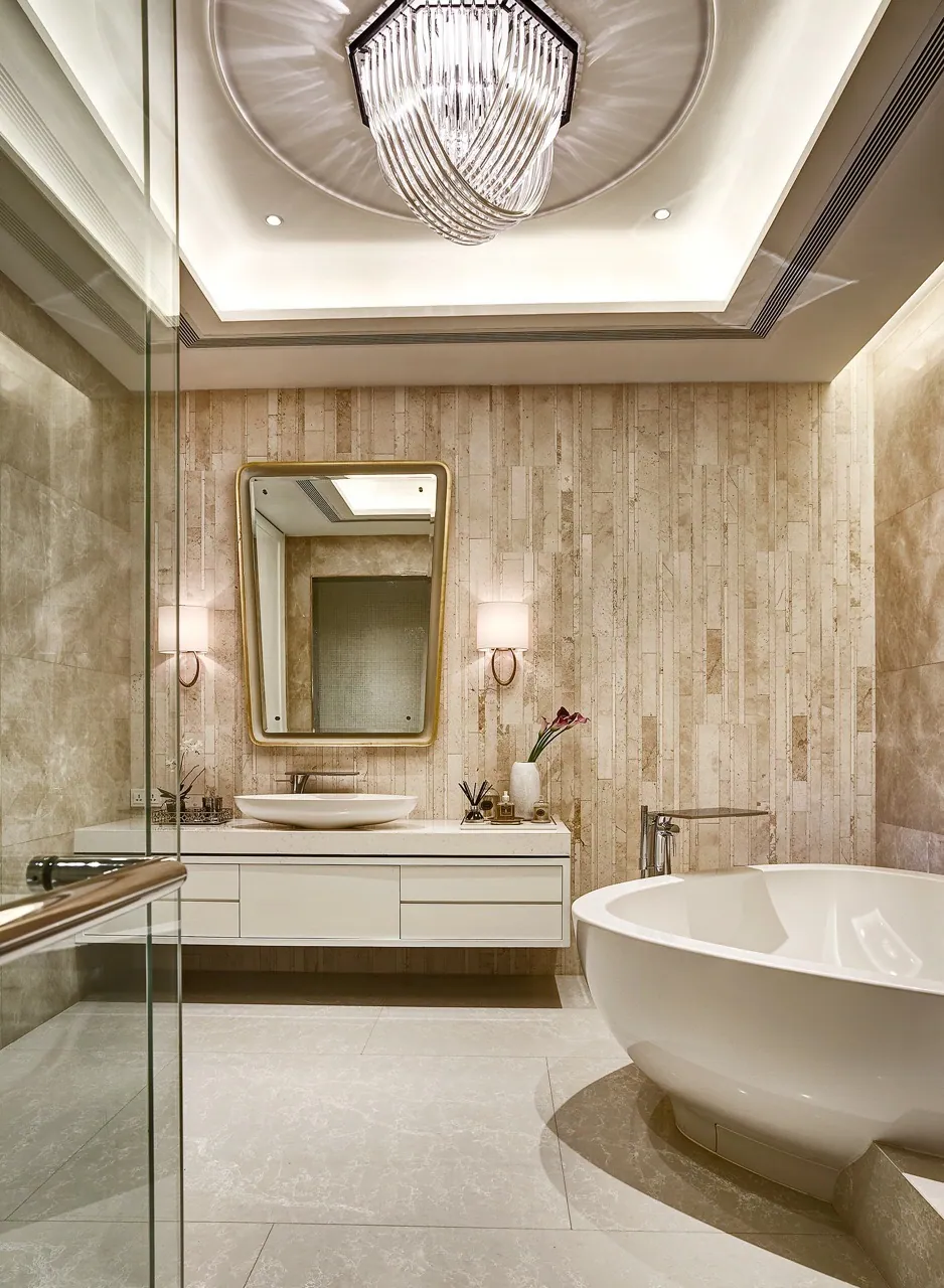 Warm bathroom at Baan Krungthep Kritha — a freestanding tub beside a travertine feature wall, with a brass-framed mirror, twin sconces and a crystal pendant in a domed ceiling recess.
