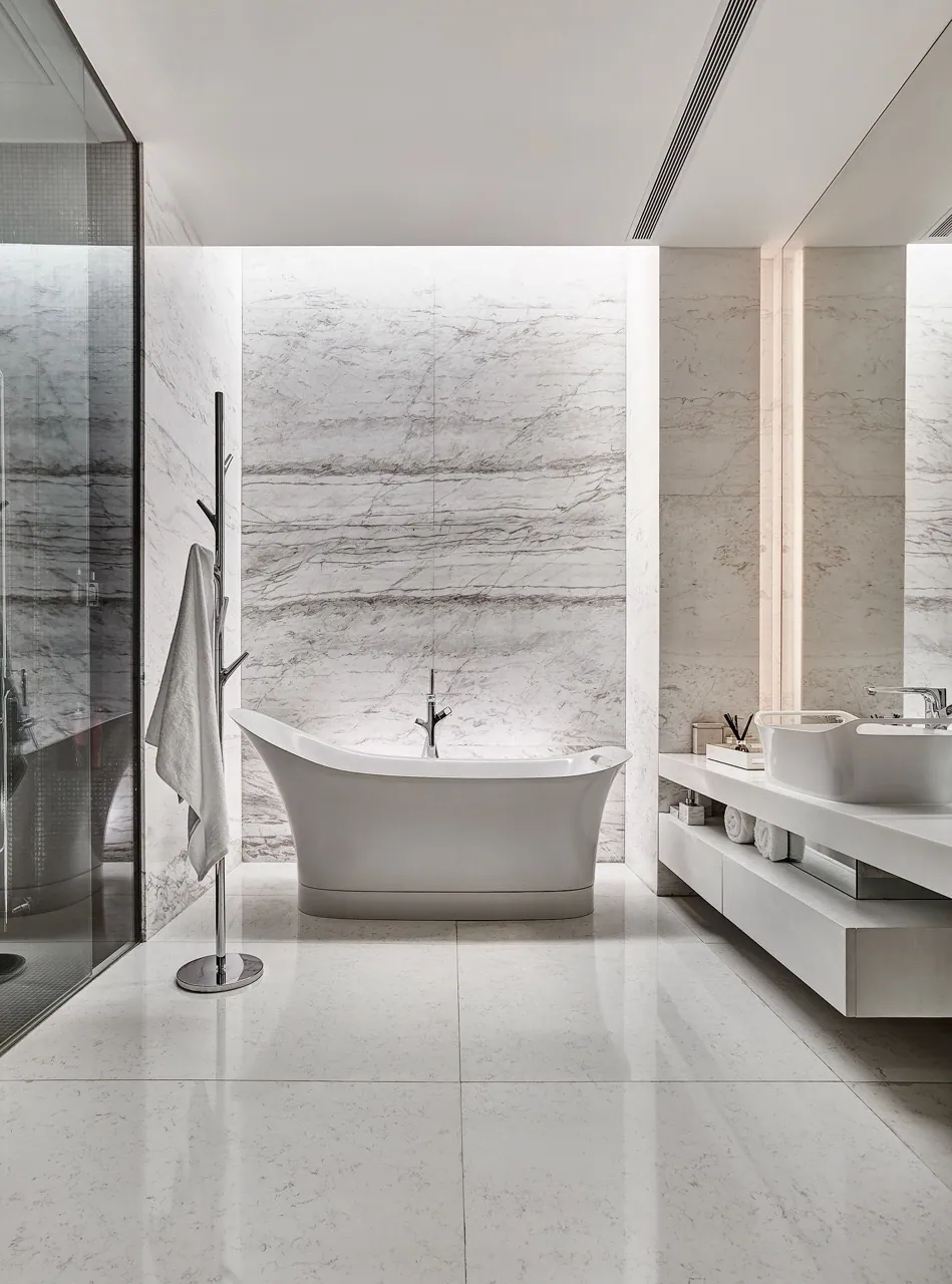 Cool bathroom at Baan Krungthep Kritha — a freestanding tub against a full-height book-matched white quartzite panel, with a floating white vanity and a glazed shower.