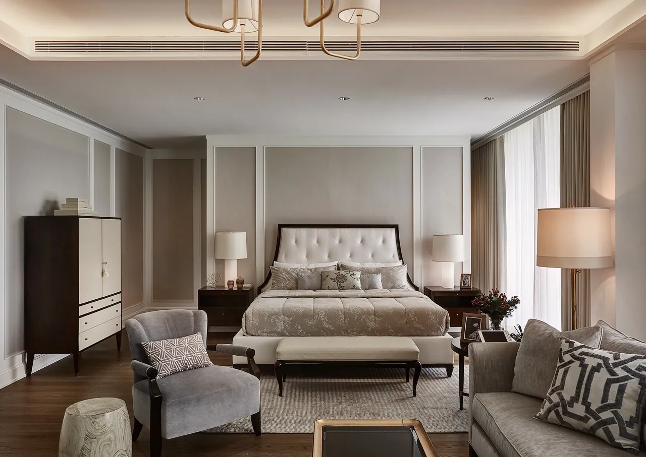 Second bedroom at Baan Krungthep Kritha — a dark-framed tufted bed set against moulded greige panelling beneath a brass ring fitting, with a velvet armchair and sofa.