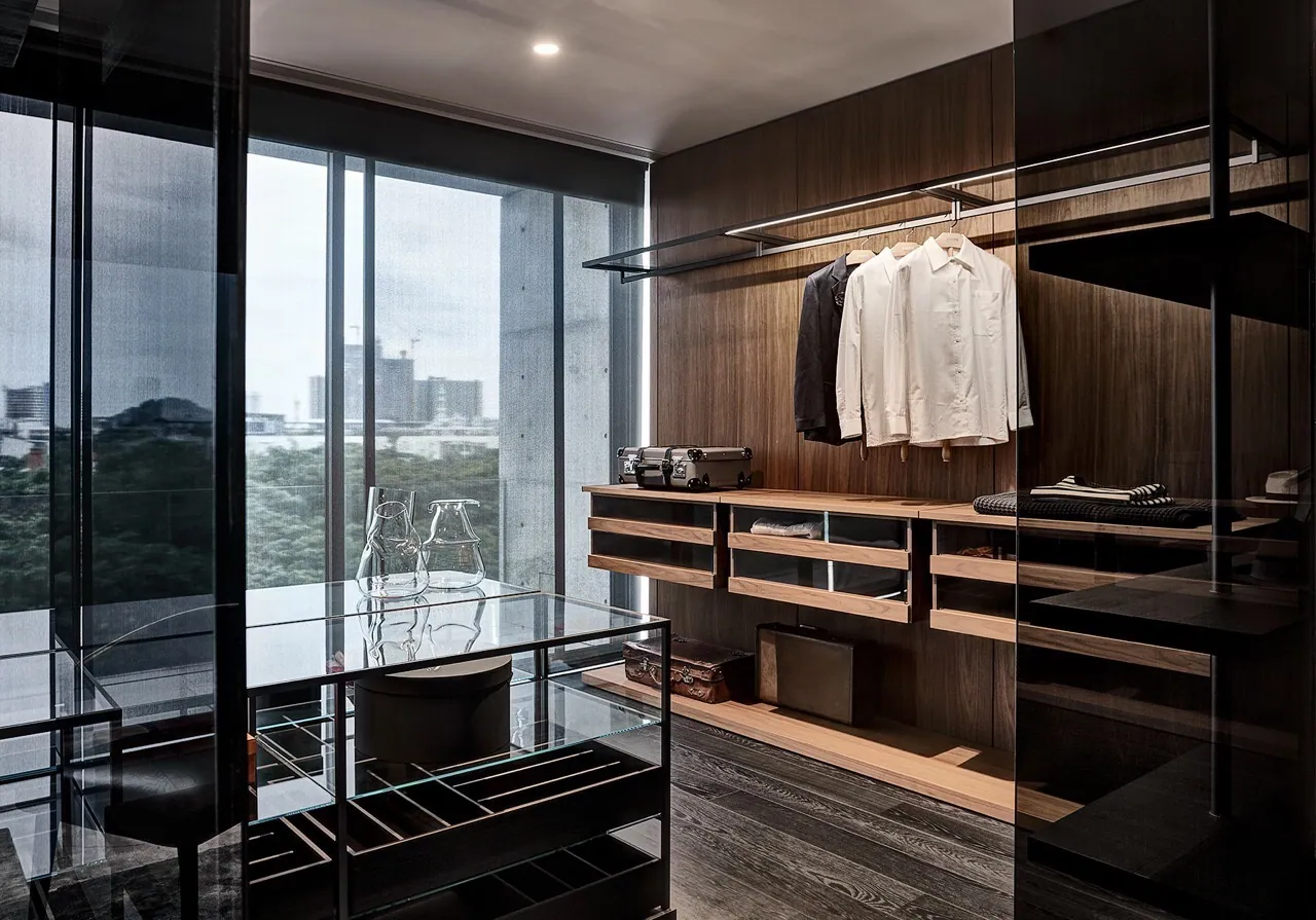 Dressing room at Baan Krungthep Kritha — walnut hanging rails and open drawers with integrated lighting, a glass-topped vitrine island, and black-framed glazing onto board-formed concrete and the skyline.