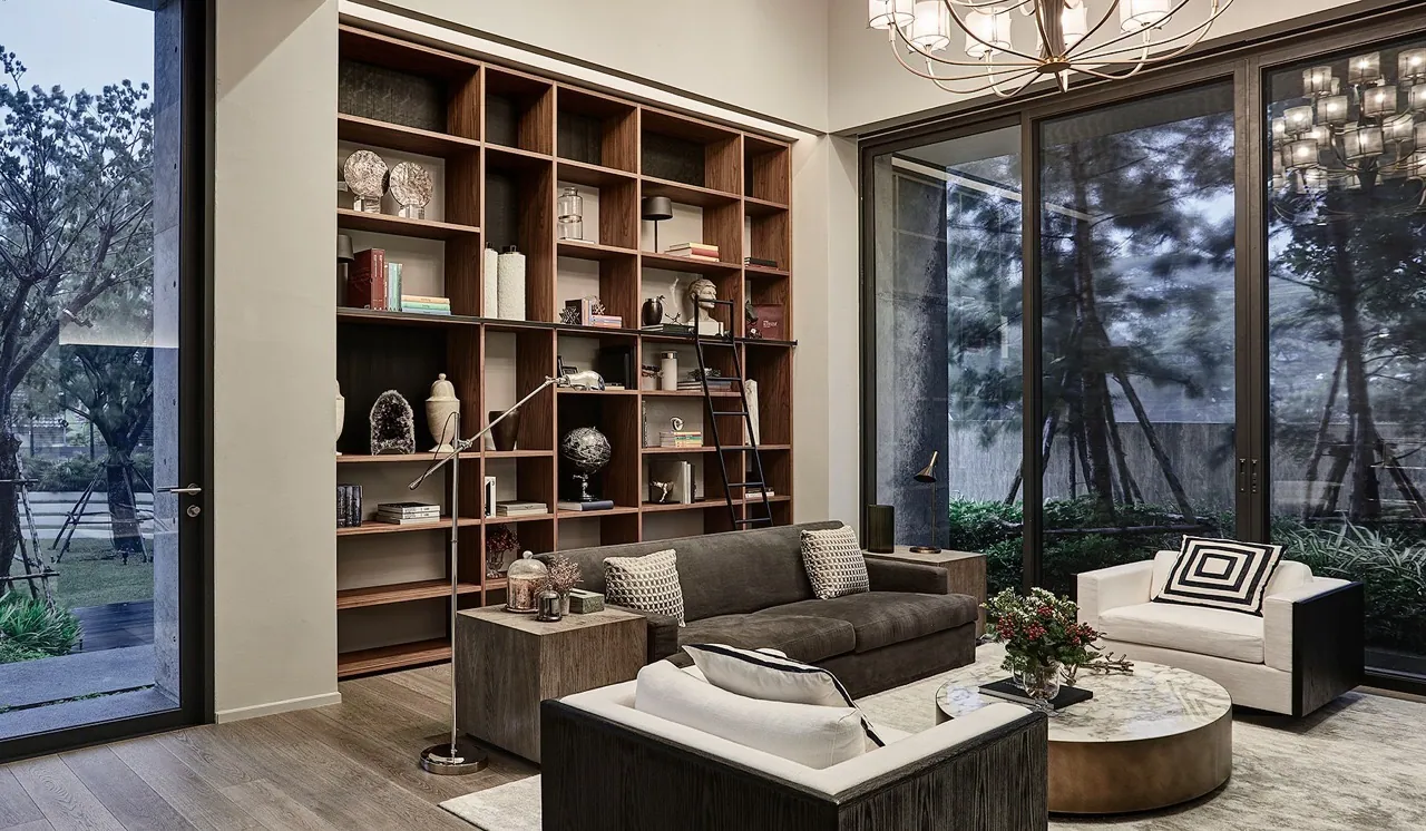 Library corner at Baan Krungthep Kritha — a full-height walnut grid bookcase with a black rolling ladder, brass ring chandelier, and glazing onto a courtyard of pines.