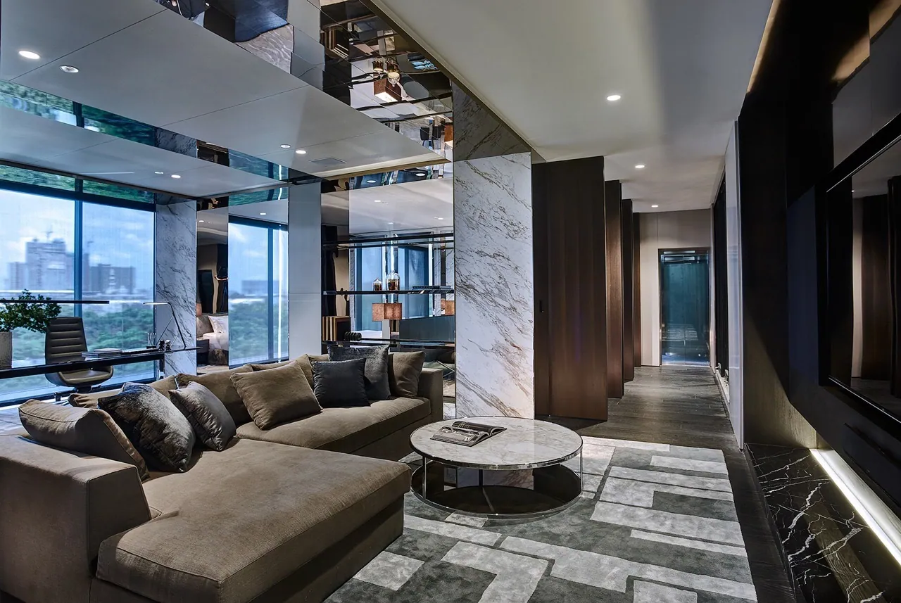 Study lounge at Baan Krungthep Kritha — a book-matched marble pier and mirrored ceiling soffit beside a deep taupe sectional, with Bangkok's skyline through full-height glazing.