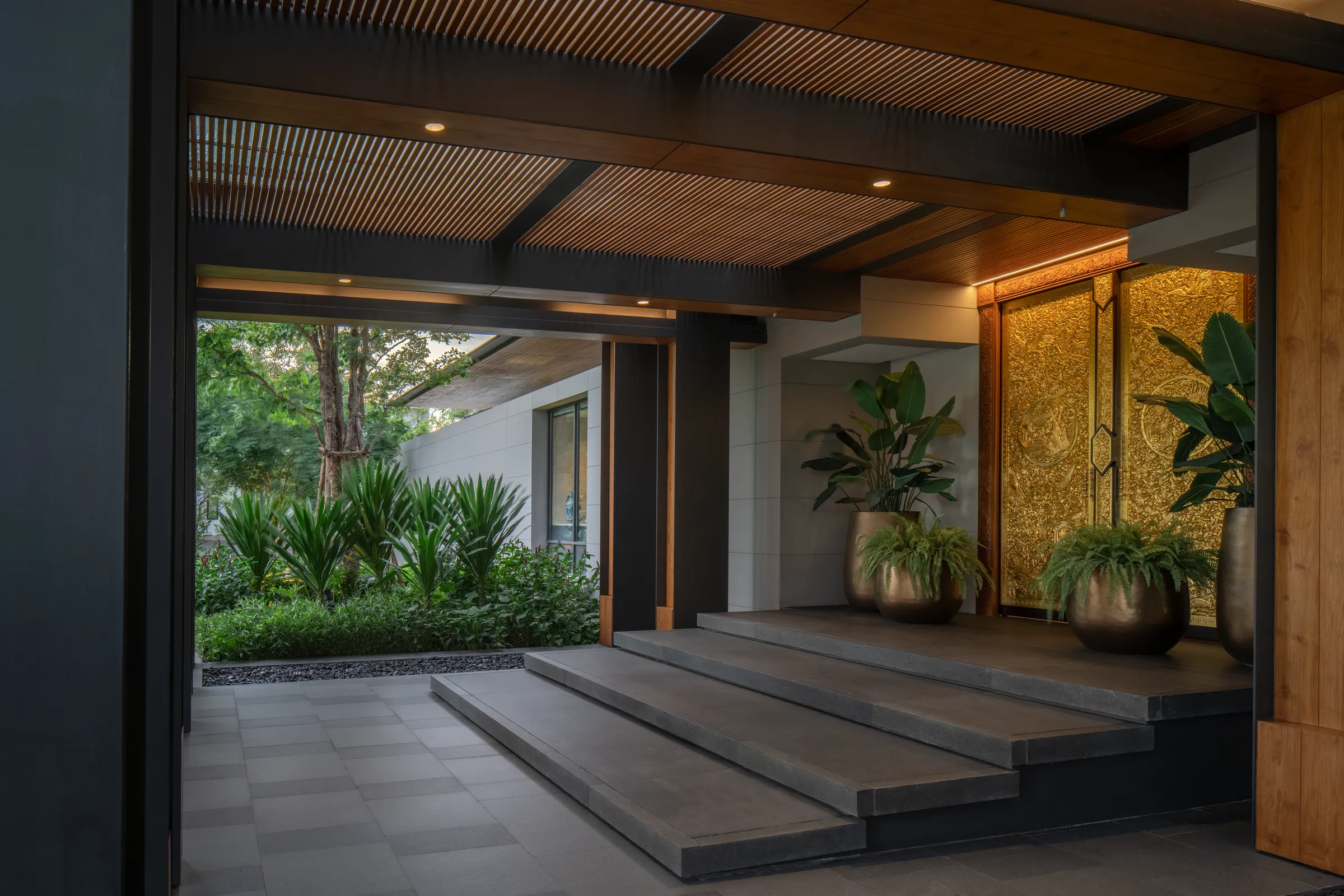 Wooden entrance portal of Khon Kaen Residence flanked by tropical plants in late-afternoon light.