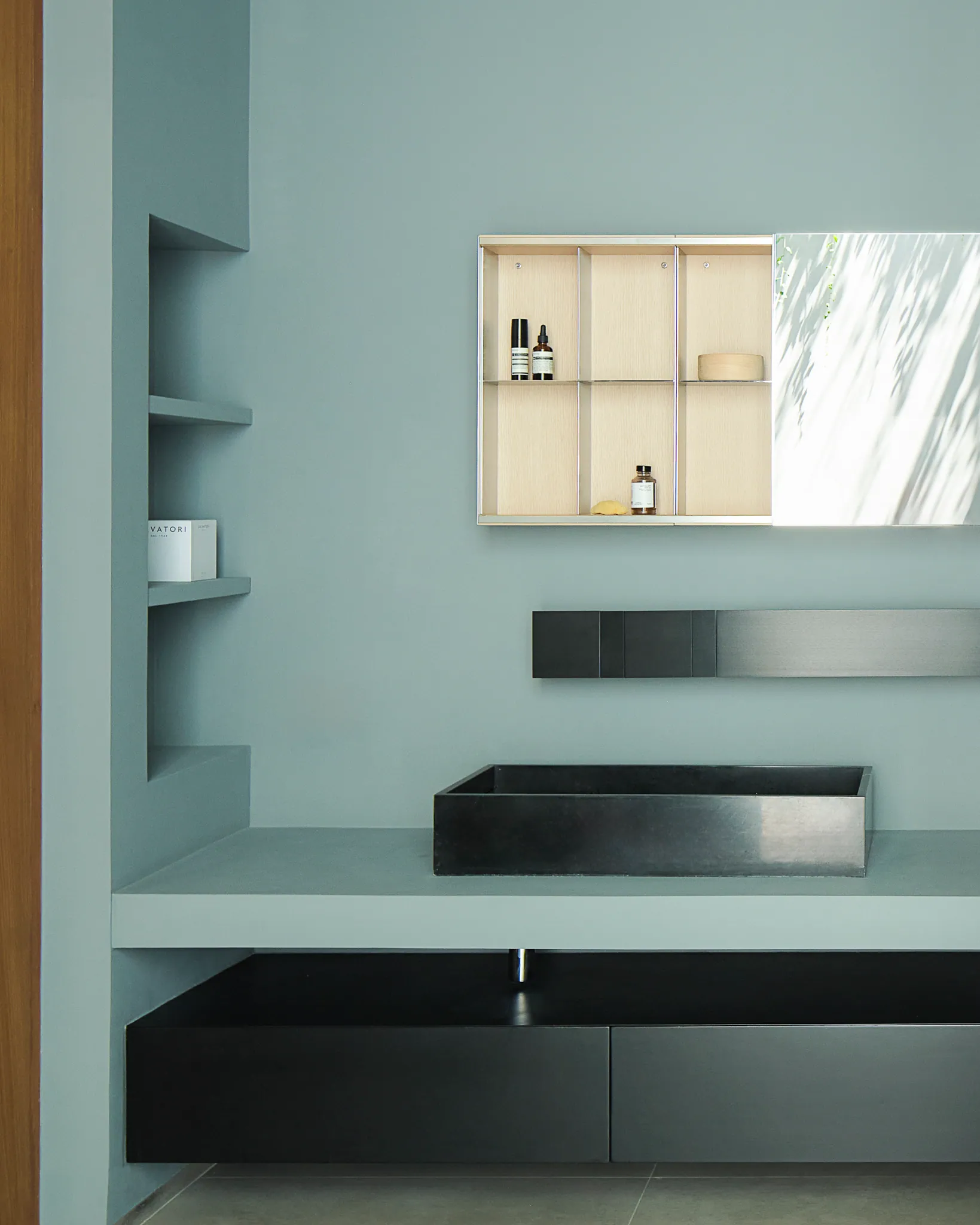 Night-mode bathroom at Black Box Project — teal-slate shelving, minimalist sink, sculptural textured vase.