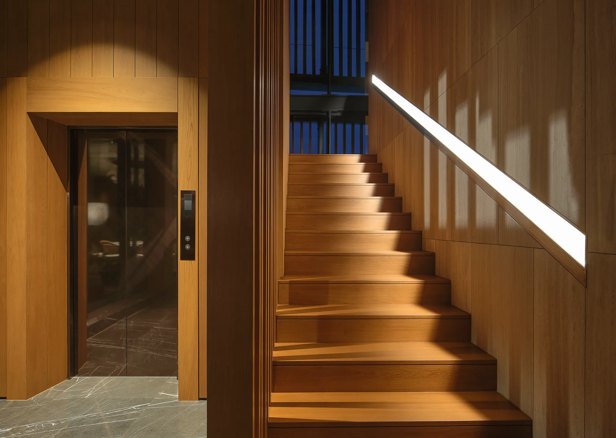 Honey-wood staircase at Black Box Project with a glowing white LED handrail under golden-hour light.