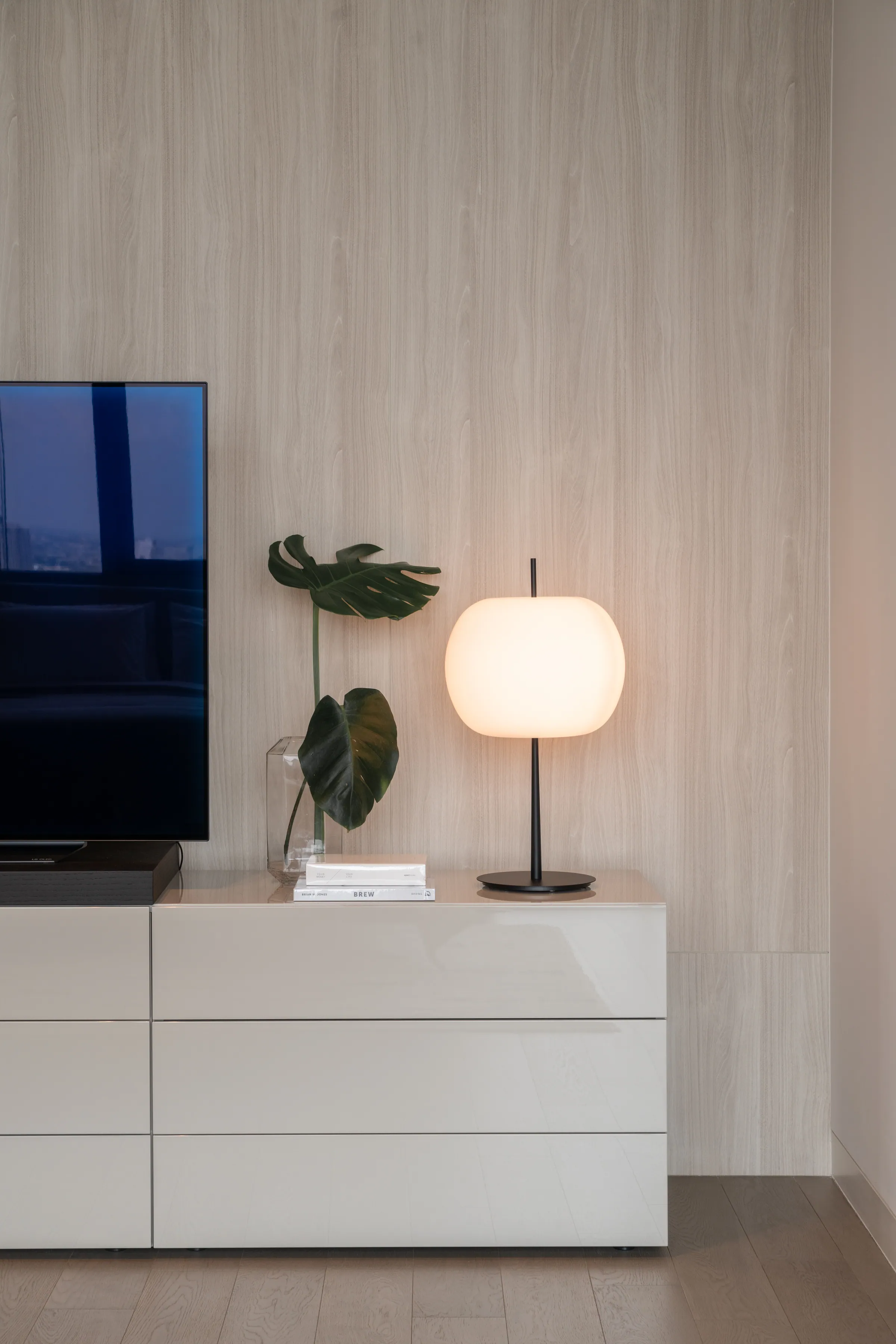 Guest bedroom at Madison Penthouse — wall-mounted screen on a pale travertine wall above a lacquer console, a globe lamp and a single monstera leaf.