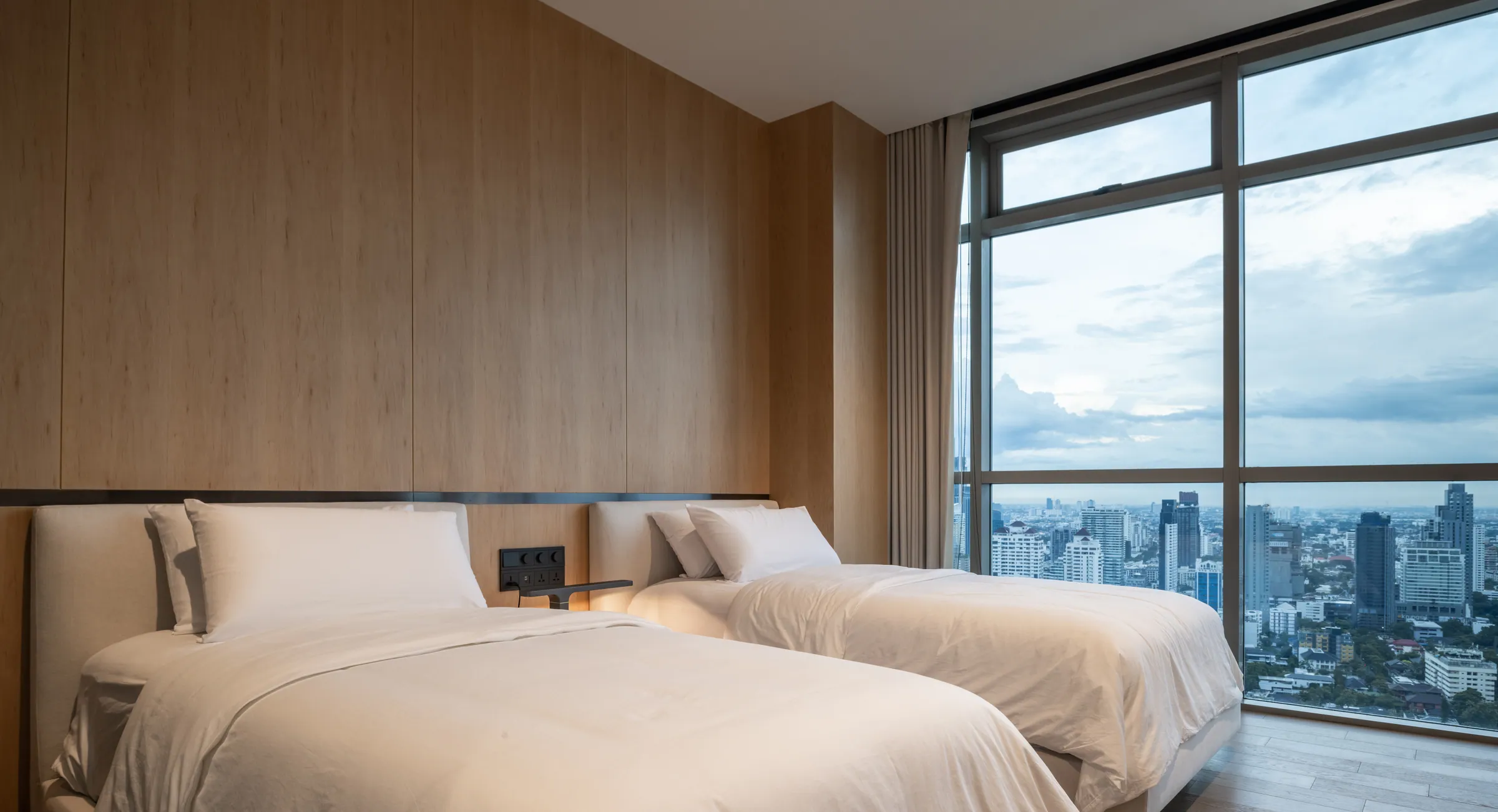 Twin bedroom at Madison Penthouse — oak headboard wall and full-height wardrobe against corner glazing over the city.