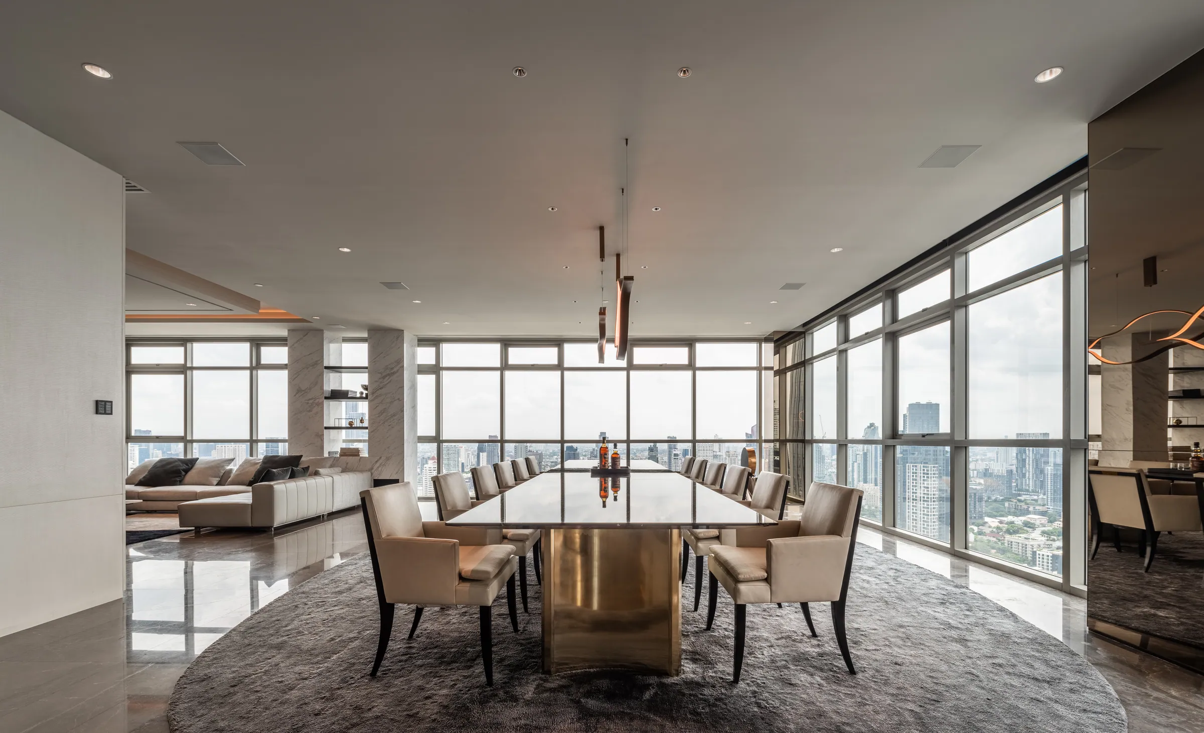 Dining room at Madison Penthouse — ten upholstered chairs around a bronze-pedestal table beneath a linear sculptural pendant, city glazing along the long wall.