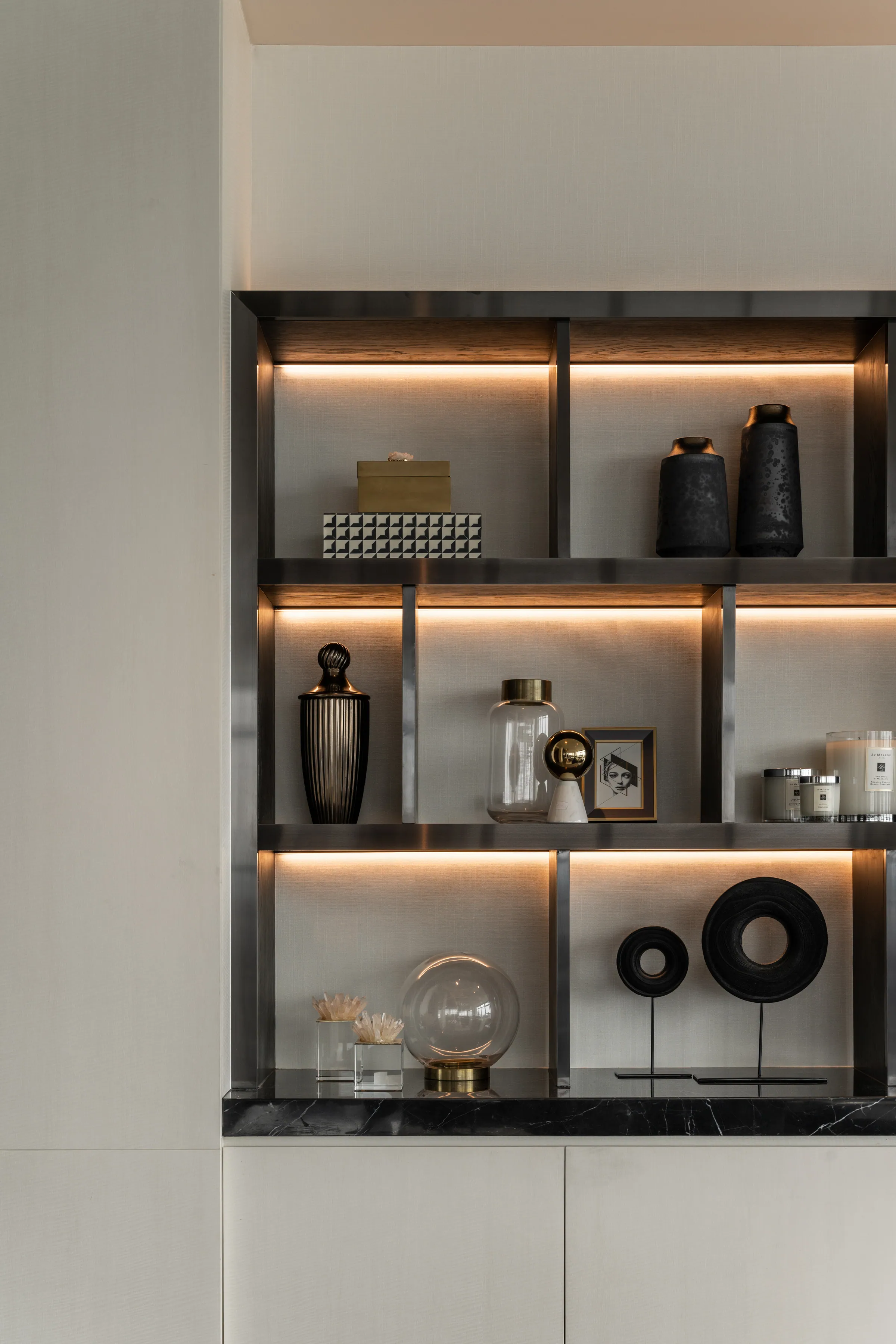 Backlit display shelving at Madison Penthouse — dark ceramic vessels, a brass box, framed photograph and candles on warm-lit ledges.