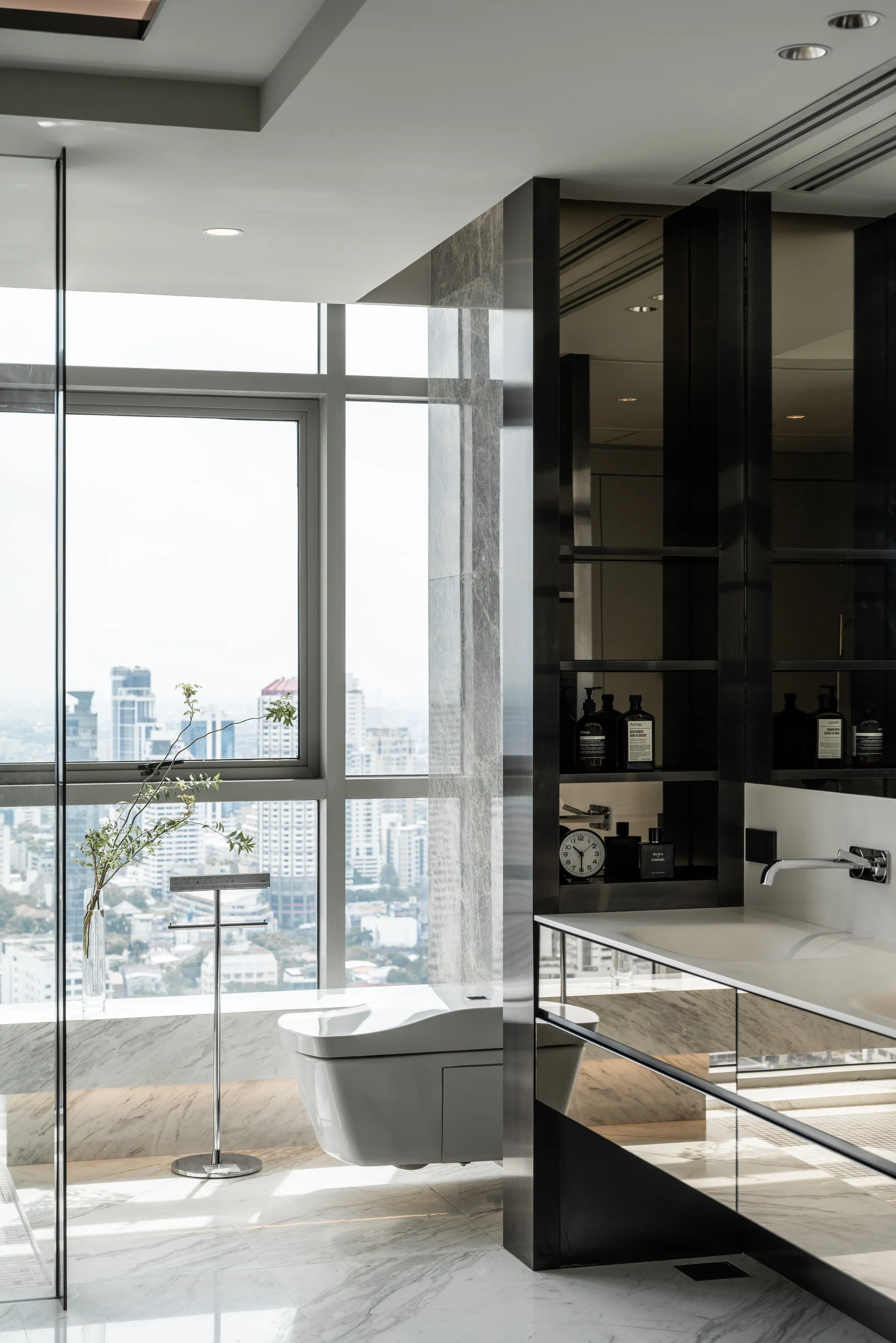 Master bathroom vanity at Madison Penthouse — wall-mounted spout over a stone counter, blackened shelving and a branch arrangement against the city window.
