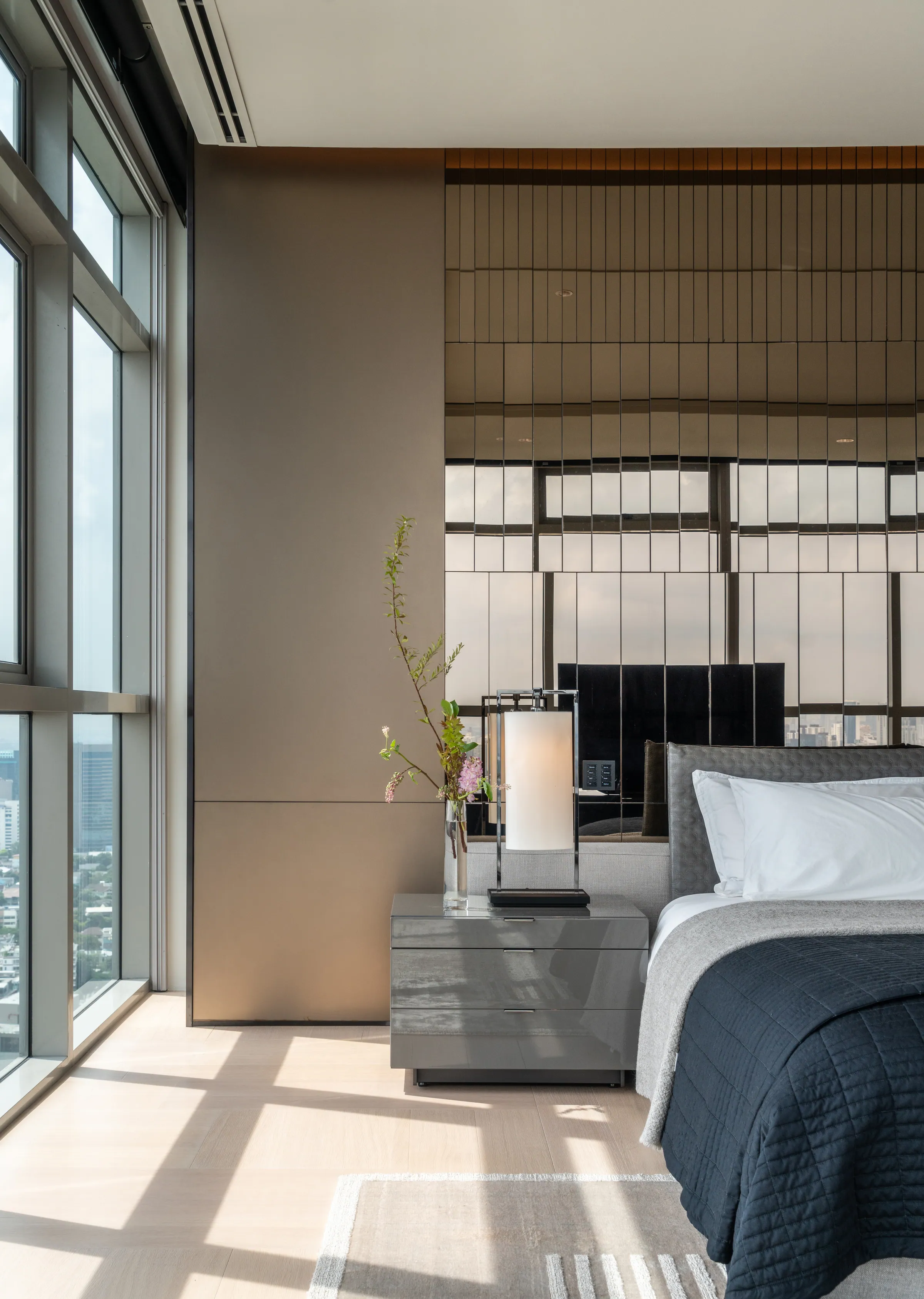 Master bedroom detail at Madison Penthouse — faceted mirrored panel wall behind a black upholstered headboard, a lantern lamp and a branch of blossom.