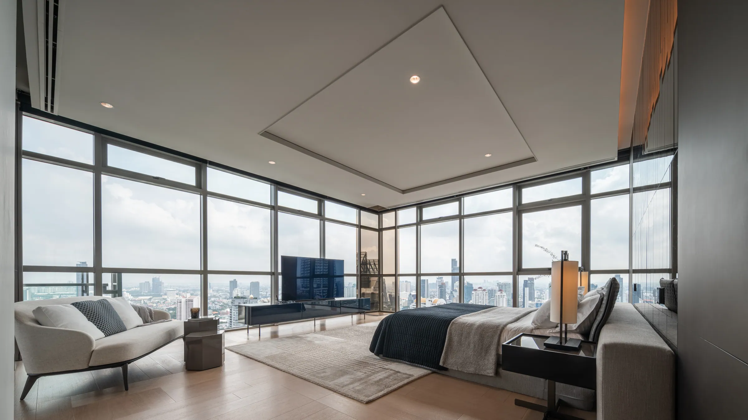 Master bedroom at Madison Penthouse — corner glazing on two sides, a low bed, chaise and media console under a recessed ceiling tray.