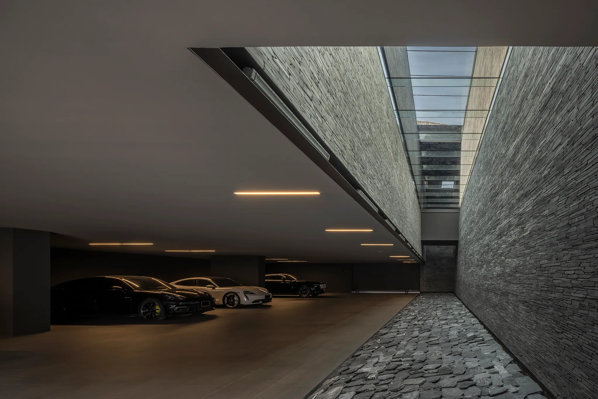 Approach to the garage at Water House — warm linear ceiling lights, textured brick wall, vehicles barely visible in shadow.